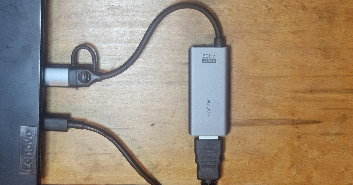 A USB HDMI capture device on a desk, connected to a laptop over USB with an HDMI cable entering from below.
